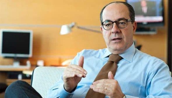 Businessman wearing a light blue shirt and glasses, gesturing while seated in an office environment. - Olive Oil Times