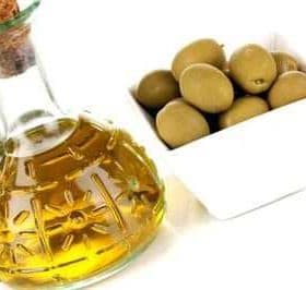 A glass bottle of olive oil with a cork stopper next to a bowl of green olives. - Olive Oil Times