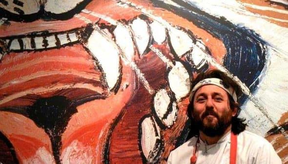 Chef with a beard and white chef's coat standing beside a large colorful mural of a lion's face. - Olive Oil Times