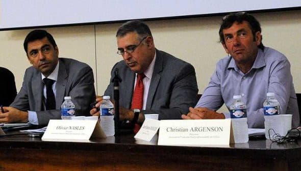 Three men seated at a conference table with water bottles and nameplates in front of them. - Olive Oil Times