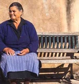 Older woman wearing a blue sweater and skirt sitting on a wooden bench against a wall. - Olive Oil Times