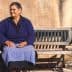 Older woman wearing a blue sweater and skirt sitting on a wooden bench against a wall. - Olive Oil Times