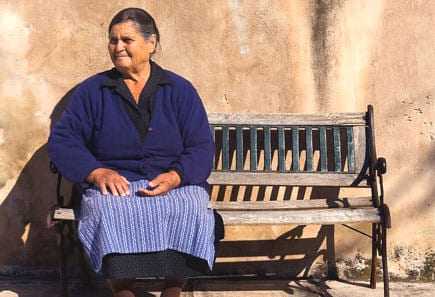 Older woman wearing a blue sweater and skirt sitting on a wooden bench against a wall. - Olive Oil Times