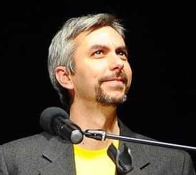 A man with gray hair and a goatee speaking into a microphone while wearing a black jacket and yellow shirt. - Olive Oil Times