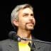 A man with gray hair and a goatee speaking into a microphone while wearing a black jacket and yellow shirt. - Olive Oil Times