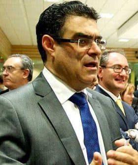 A man in a gray suit with a blue tie speaking in a group setting with other men in suits. - Olive Oil Times