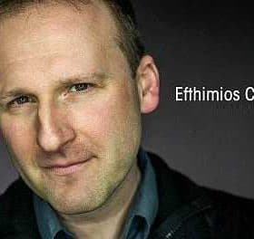 Close-up portrait of a man with short hair and a slight smile against a dark background. - Olive Oil Times