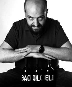 Man with a beard sitting in front of three bottles labeled 'Bacchus' in black and white. - Olive Oil Times