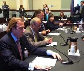 A group of individuals seated at a conference table during a meeting with documents and a coffee pot visible. - Olive Oil Times