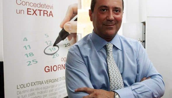 A businessman in a blue dress shirt standing with arms crossed in an office environment. - Olive Oil Times