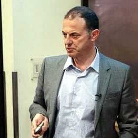 Man wearing a gray suit and holding a pointer while presenting in an indoor setting. - Olive Oil Times