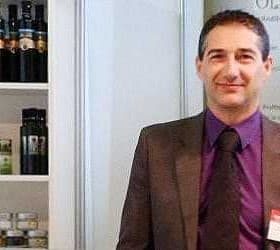 A man in a suit standing in front of a display of olive oil products on shelves. - Olive Oil Times
