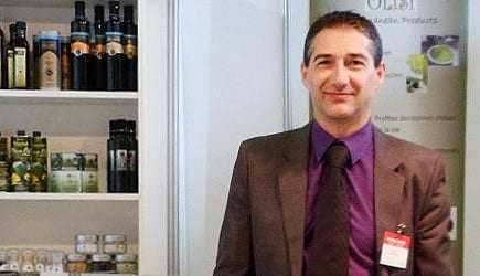 A man in a suit standing in front of a display of olive oil products on shelves. - Olive Oil Times