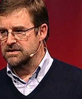 A man wearing glasses and a sweater speaking into a microphone at a conference. - Olive Oil Times