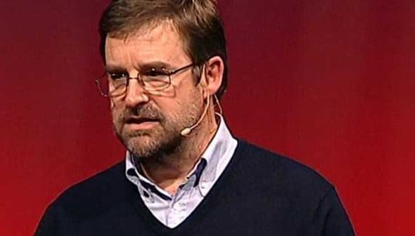 A man wearing glasses and a sweater speaking into a microphone at a conference. - Olive Oil Times