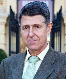 A man wearing a brown suit and a light green tie, standing outdoors in front of a building. - Olive Oil Times