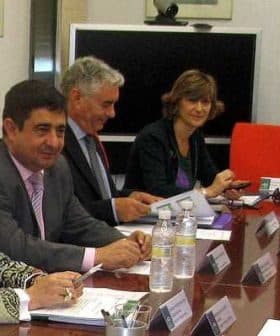 Group of individuals seated at a conference table during a meeting, with documents and water bottles present. - Olive Oil Times