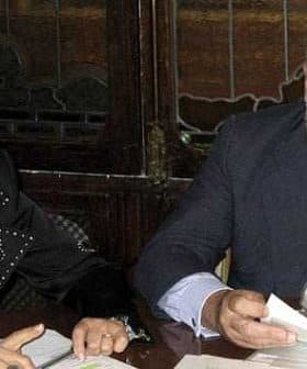 Two individuals seated at a table, one reviewing documents while the other observes attentively. - Olive Oil Times