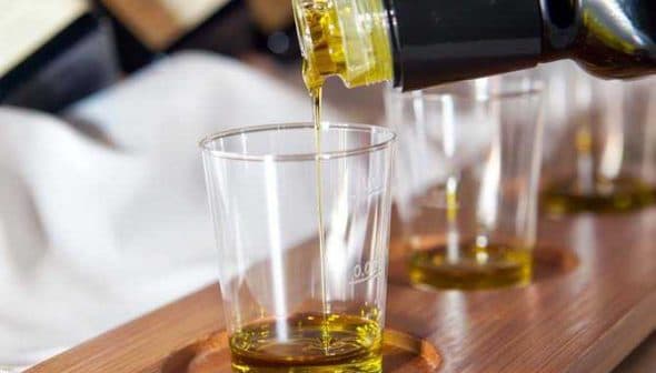 Olive oil being poured from a bottle into a clear glass sample cup on a wooden tray. - Olive Oil Times
