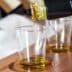 Olive oil being poured from a bottle into a clear glass sample cup on a wooden tray. - Olive Oil Times