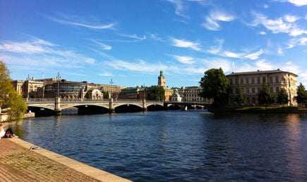 View of a bridge over water in Stockholm with buildings and trees in the background. - Olive Oil Times