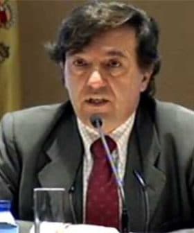 Man in a suit speaking at a conference with a Spanish flag visible in the background. - Olive Oil Times