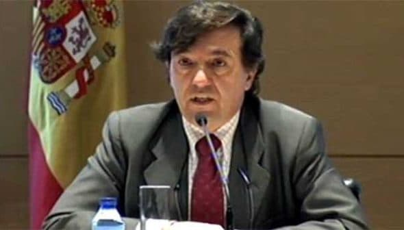 Man in a suit speaking at a conference with a Spanish flag visible in the background. - Olive Oil Times