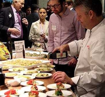 Chef pouring olive oil over a plate of food at a culinary event with attendees observing. - Olive Oil Times