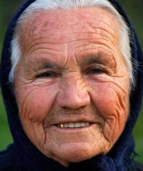 Elderly woman with a warm smile, wearing a dark headscarf and showing facial wrinkles. - Olive Oil Times
