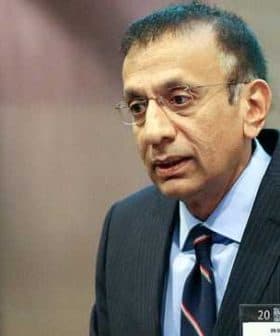 A man in a dark suit and glasses speaking during a presentation at an event. - Olive Oil Times