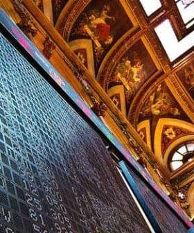 Large electronic stock market display board in a historic building with ornate ceiling. - Olive Oil Times