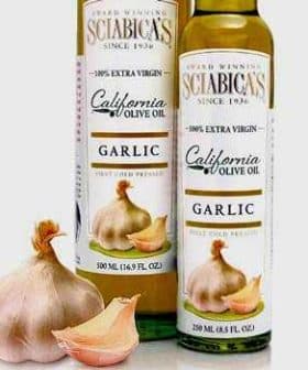 Two bottles of garlic olive oil with a garlic bulb in front on a neutral background. - Olive Oil Times