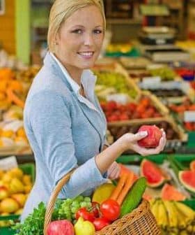 Woman in a gray blazer holding a red apple while carrying a basket of fresh fruits and vegetables. - Olive Oil Times
