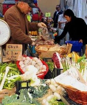 A vendor weighing vegetables at a market stall with various fresh produce displayed. - Olive Oil Times