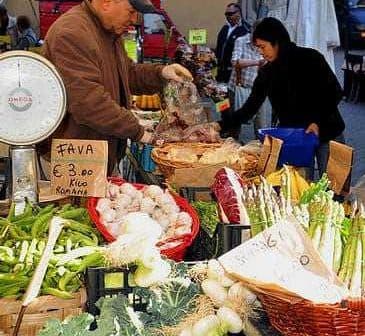 A vendor weighing vegetables at a market stall with various fresh produce displayed. - Olive Oil Times