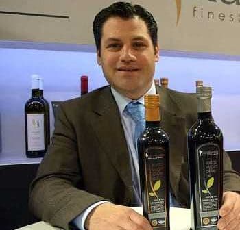 A man in a suit sitting at a table with two bottles of olive oil in front of him. - Olive Oil Times