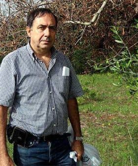 Man wearing a checkered shirt and holding bags while standing outdoors in a green area. - Olive Oil Times