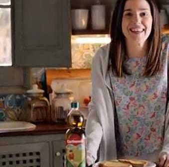 Woman smiling while holding a plate of toast next to a bottle of Carbonell olive oil in a kitchen. - Olive Oil Times