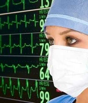 Healthcare professional in a surgical mask observing heart rate monitor with ECG readings in the background. - Olive Oil Times