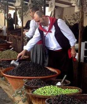 A man in traditional attire selecting olives from large bowls at a market stall. - Olive Oil Times