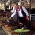 A man in traditional attire selecting olives from large bowls at a market stall. - Olive Oil Times