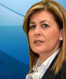 Woman with light brown hair wearing a dark blazer and white shirt against a blue background. - Olive Oil Times