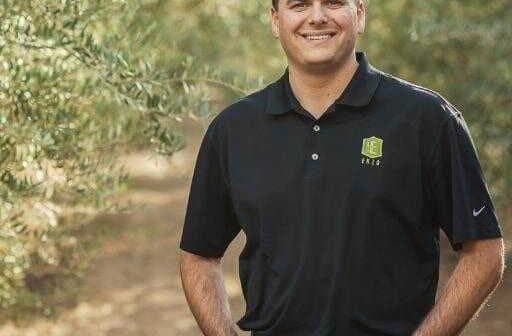 Man wearing a black polo shirt and jeans standing in an olive grove with trees in the background. - Olive Oil Times