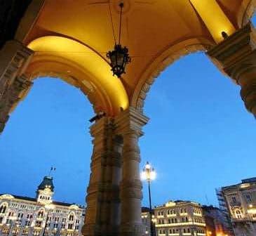 Archway with decorative columns and lanterns, overlooking a city square in Trieste during twilight. - Olive Oil Times