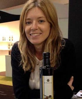 Woman with long blonde hair smiling while holding a bottle of olive oil. - Olive Oil Times