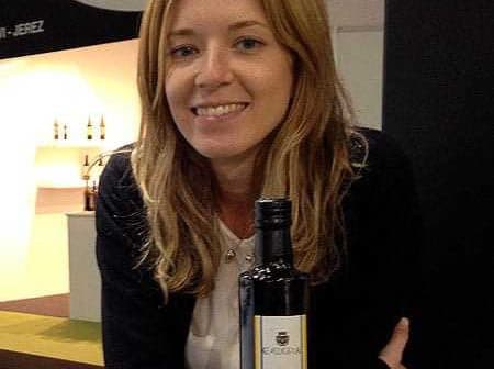 Woman with long blonde hair smiling while holding a bottle of olive oil. - Olive Oil Times