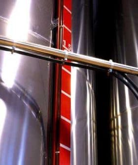 Close-up of a stainless steel storage tank with a measurement scale and piping. - Olive Oil Times