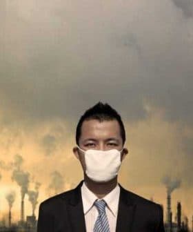 Businessman in a suit and mask standing against an industrial backdrop with smoke stacks and cloudy sky. - Olive Oil Times