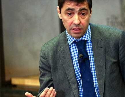 Man wearing a grey suit and blue checkered shirt speaking at an event. - Olive Oil Times