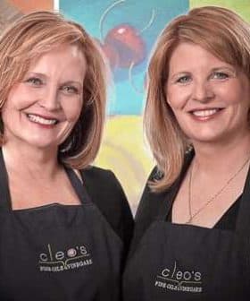 Two women wearing black aprons with 'Cleo's' logo, smiling at the camera in a studio setting. - Olive Oil Times